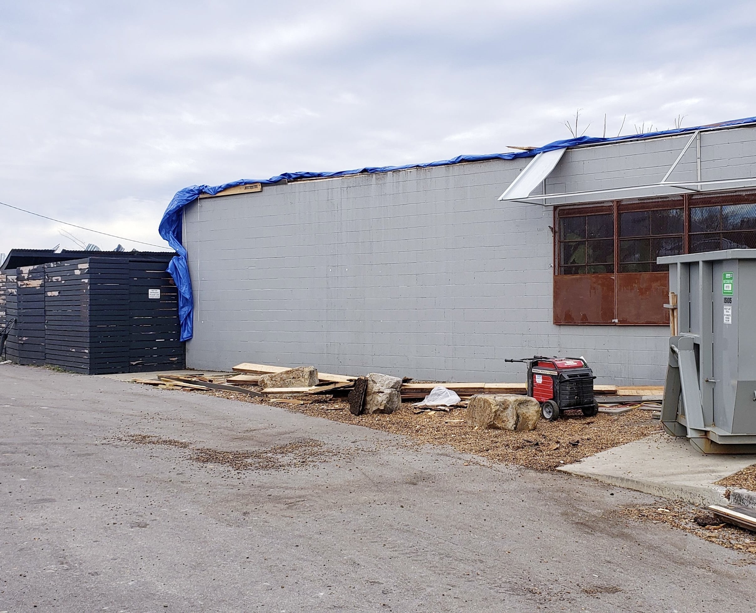 Side view of exterior tornado damage showing a gray wall with a blue tarp, piles of wood, a small generator, and a nearby dumpster.