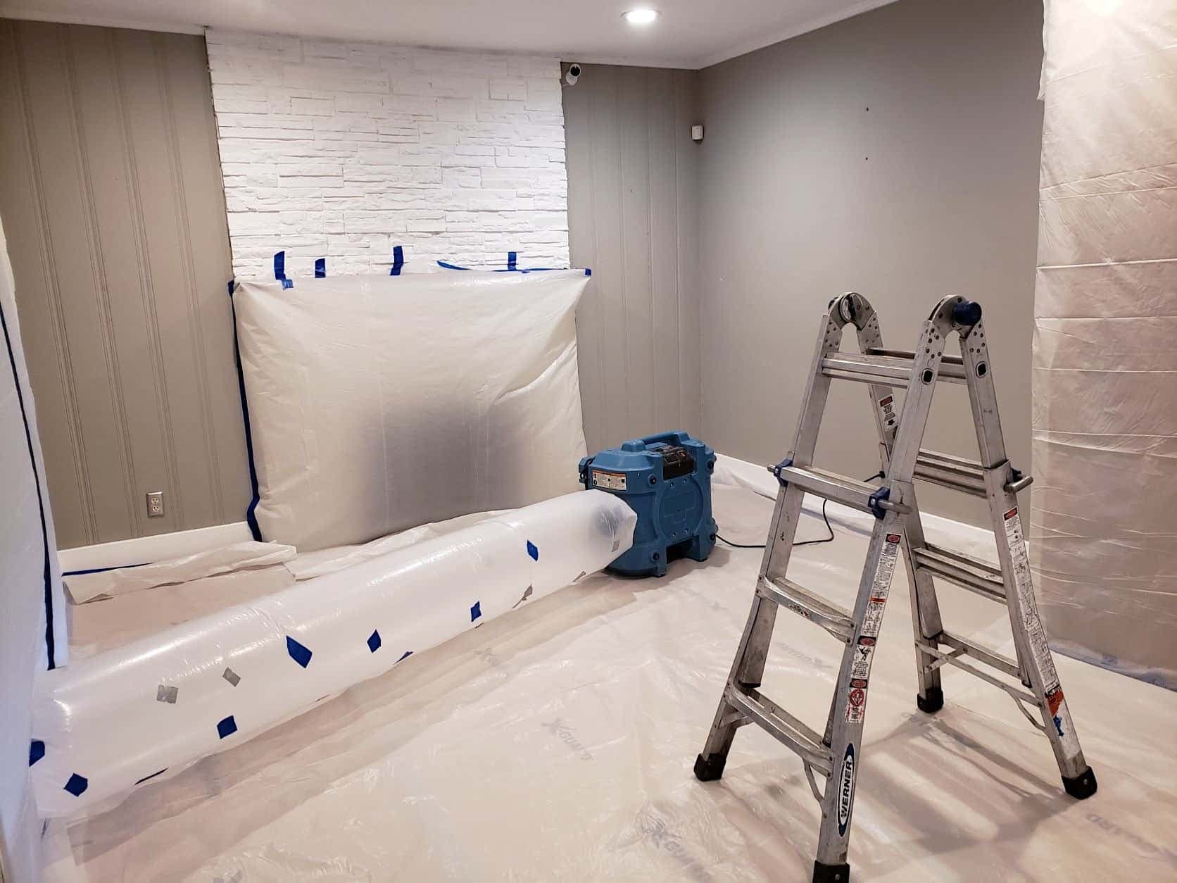 Blue air scrubber connected to plastic ducting in a room with containment sheeting during water damage restoration